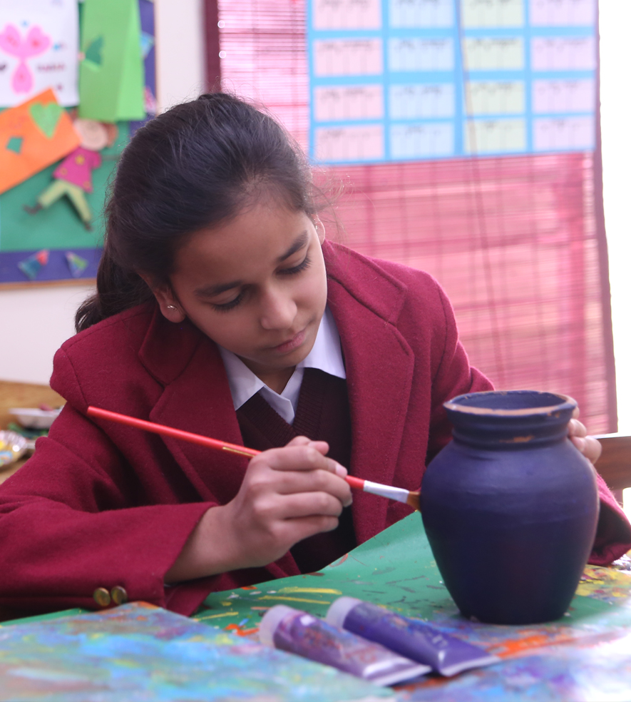In the image, there is a young student engaged in painting a piece of pottery. She appears focused and attentive to her task, holding a paintbrush and carefully applying color to the pot. The setting suggests an art class environment, which is often a space where students explore their creativity and develop their artistic skills. The school uniform implies that this activity is part of her regular school curriculum, highlighting the school's emphasis on providing a holistic education that includes artistic expression.