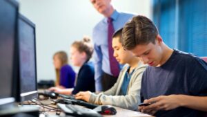 A teacher supervising a group of focused students working at computers in a school computer lab, with one student in the foreground using a smartphone.